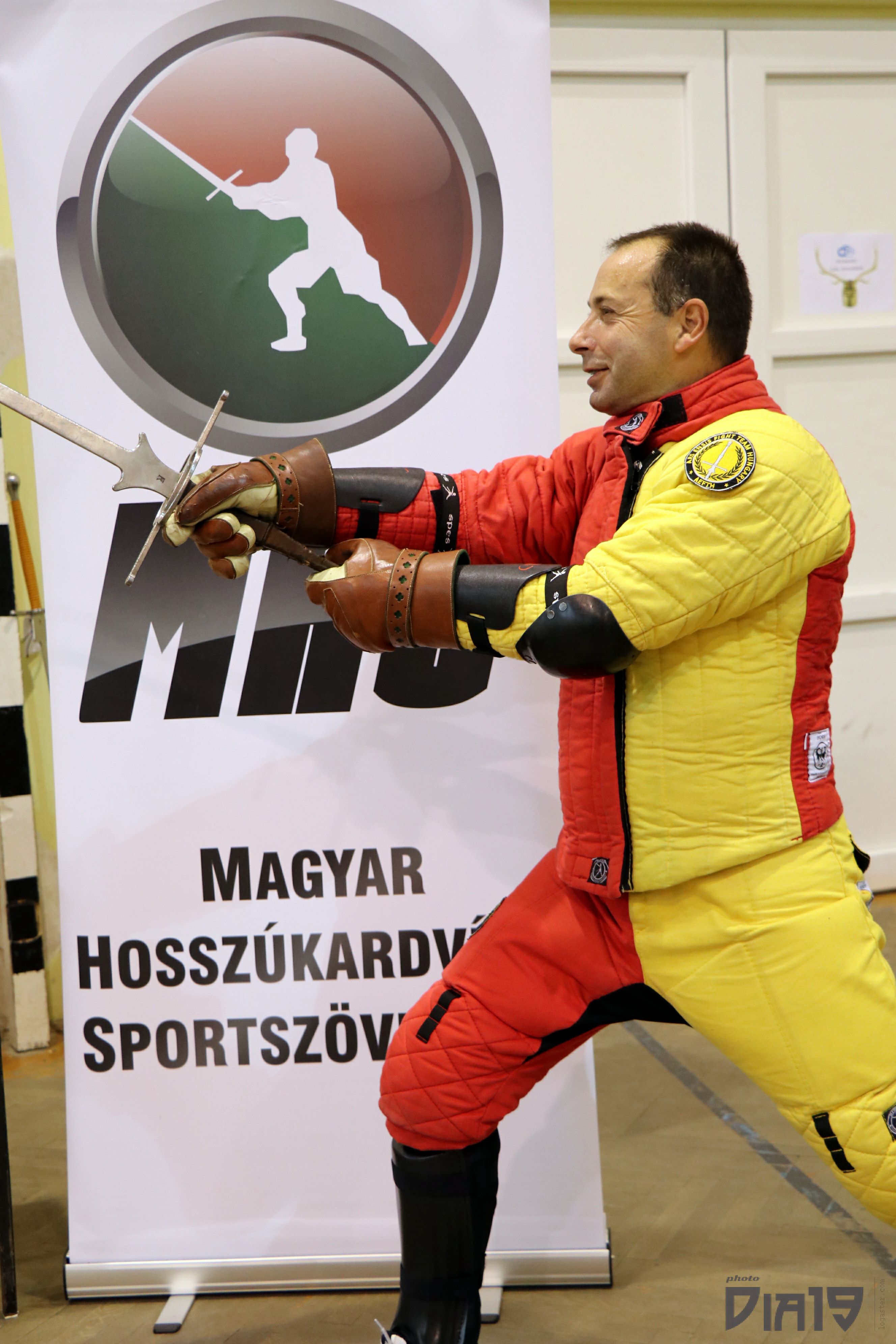 Bence László Tarr in protective HEMA equipment beside the Hungarian Longsword Association banner